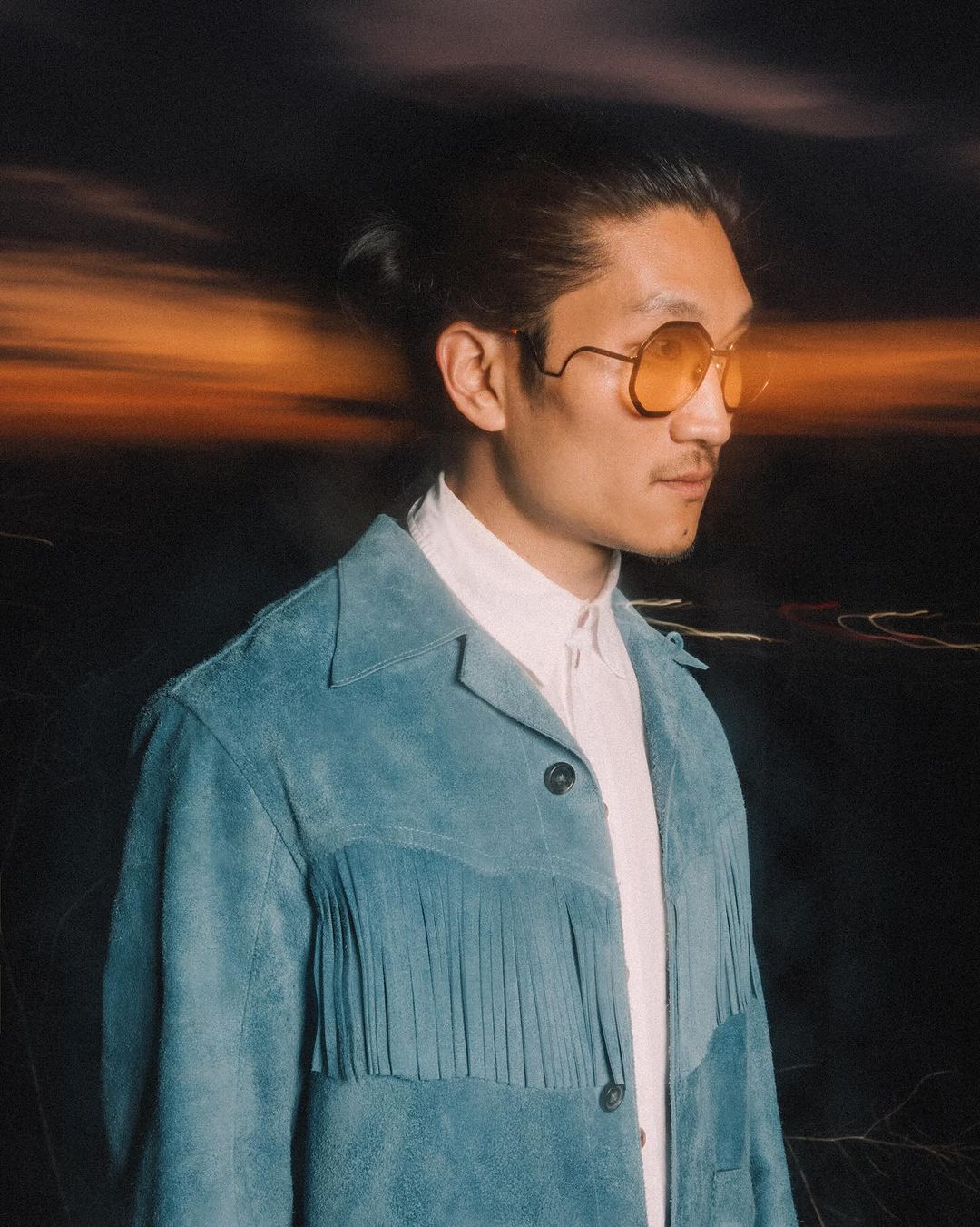 Portrait of an adult male wearing a blue fringe jacket and glasses against a blurred background with an orange sky and cityscape.