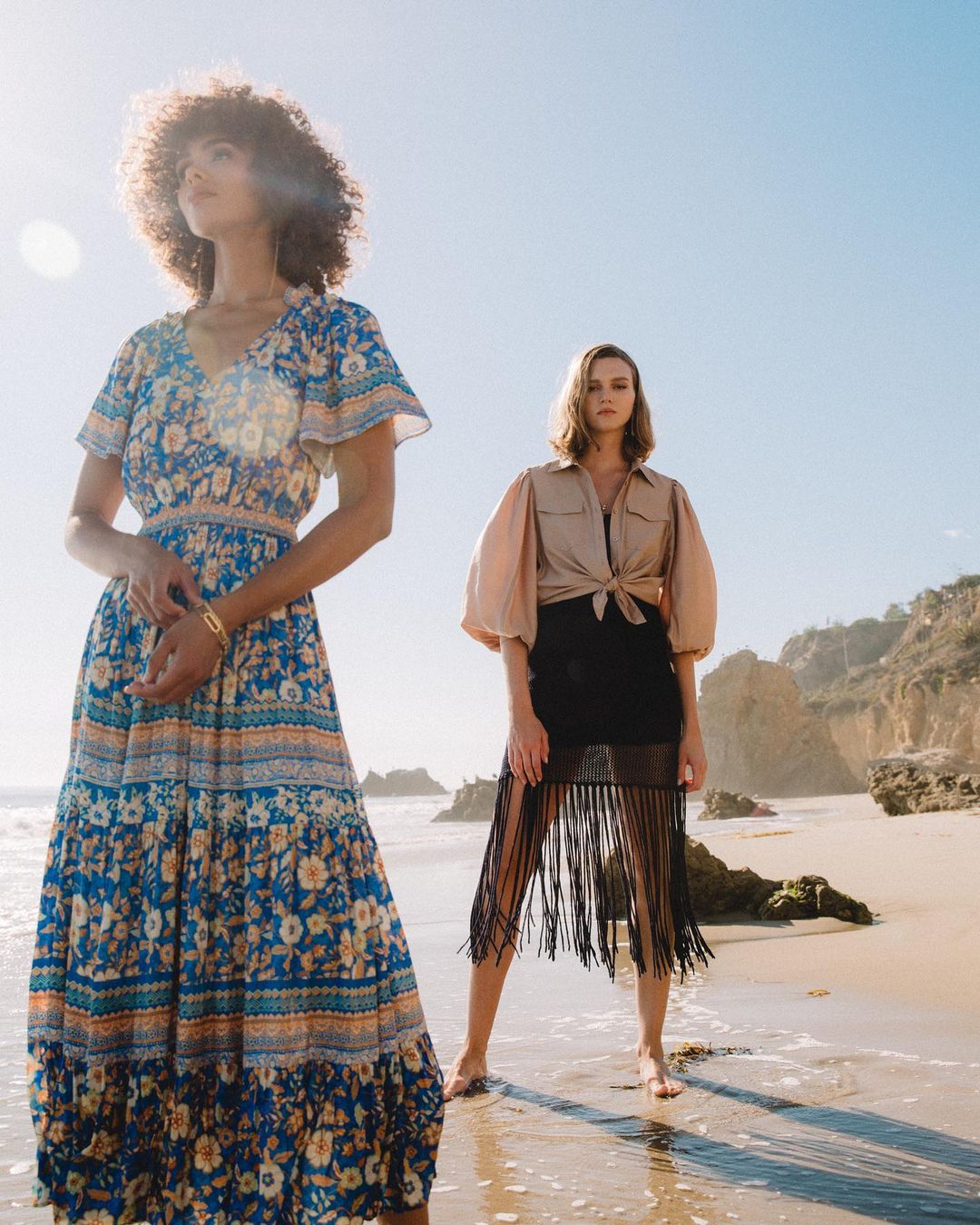 Two women pose on a sandy beach during daytime. One wears a blue floral dress, the other a beige shirt and black skirt with fringe.