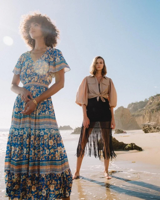 Two women pose on a sandy beach during daytime. One wears a blue floral dress, the other a beige shirt and black skirt with fringe.