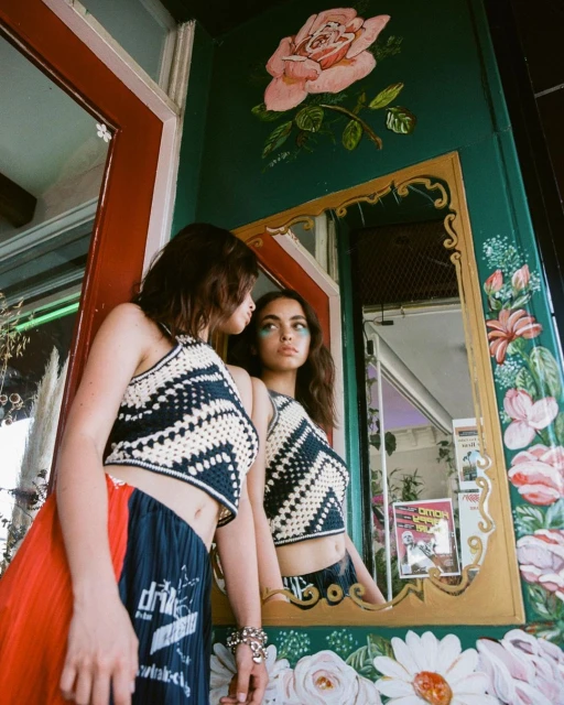 A young woman in a crocheted top and skirt poses beside a mirror with painted floral decorations in an indoor setting with natural light.