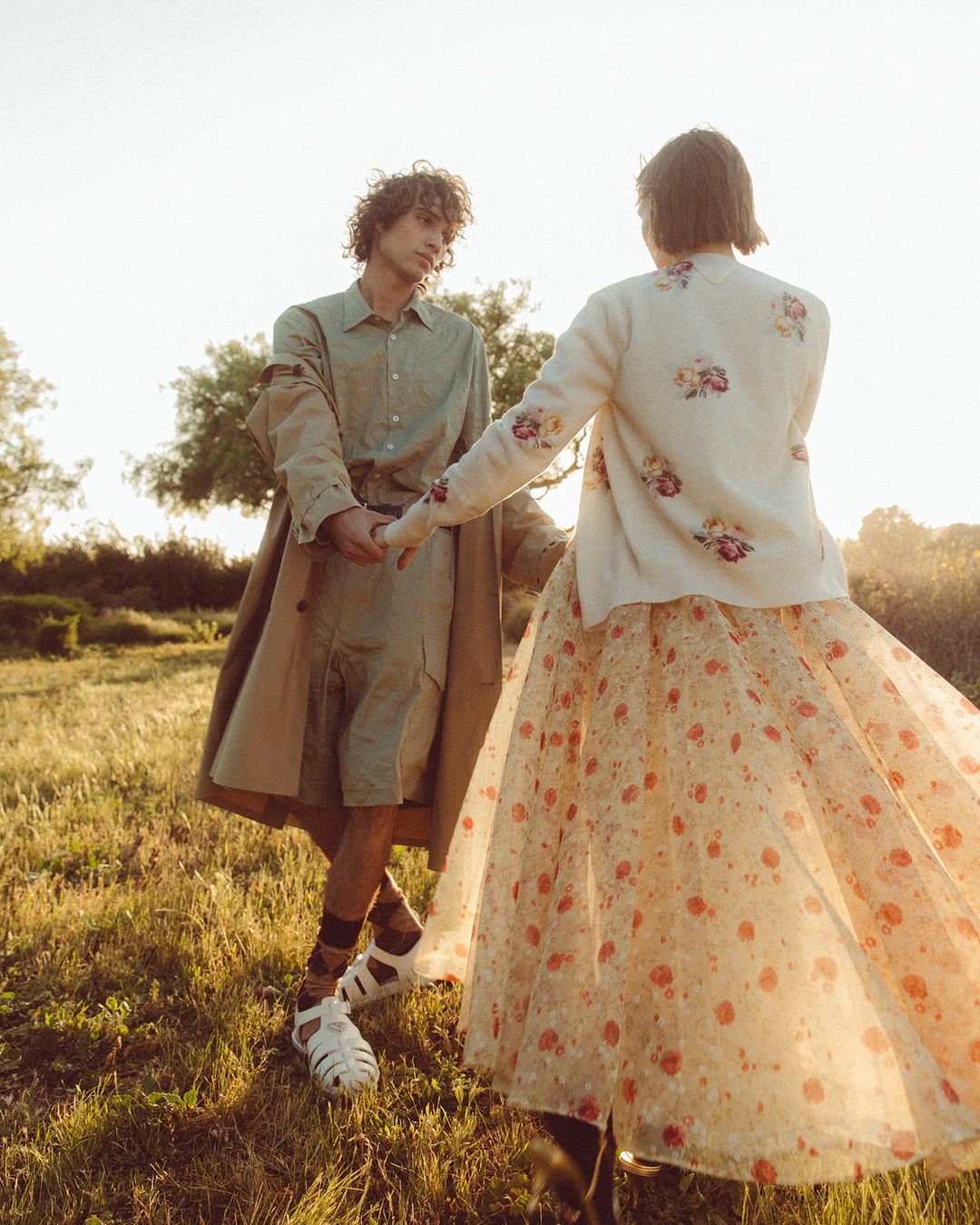 A young couple holds hands in a sunlit field. The man wears a tan overcoat and shorts, while the woman wears a floral dress, outdoors.