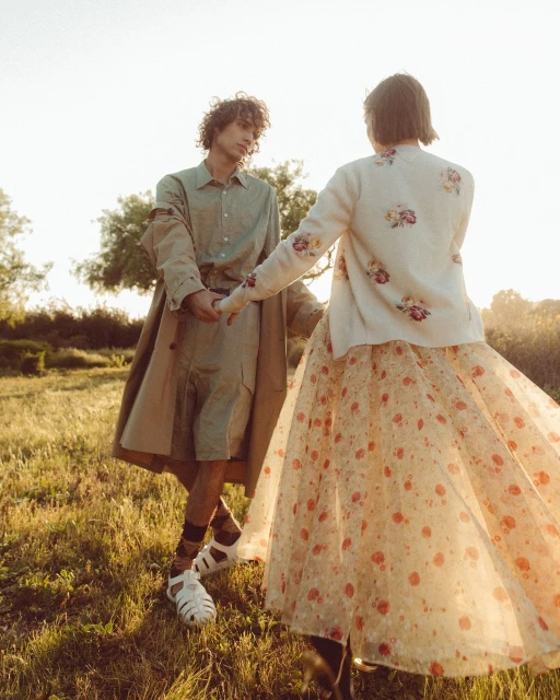 A young couple holds hands in a sunlit field. The man wears a tan overcoat and shorts, while the woman wears a floral dress, outdoors.