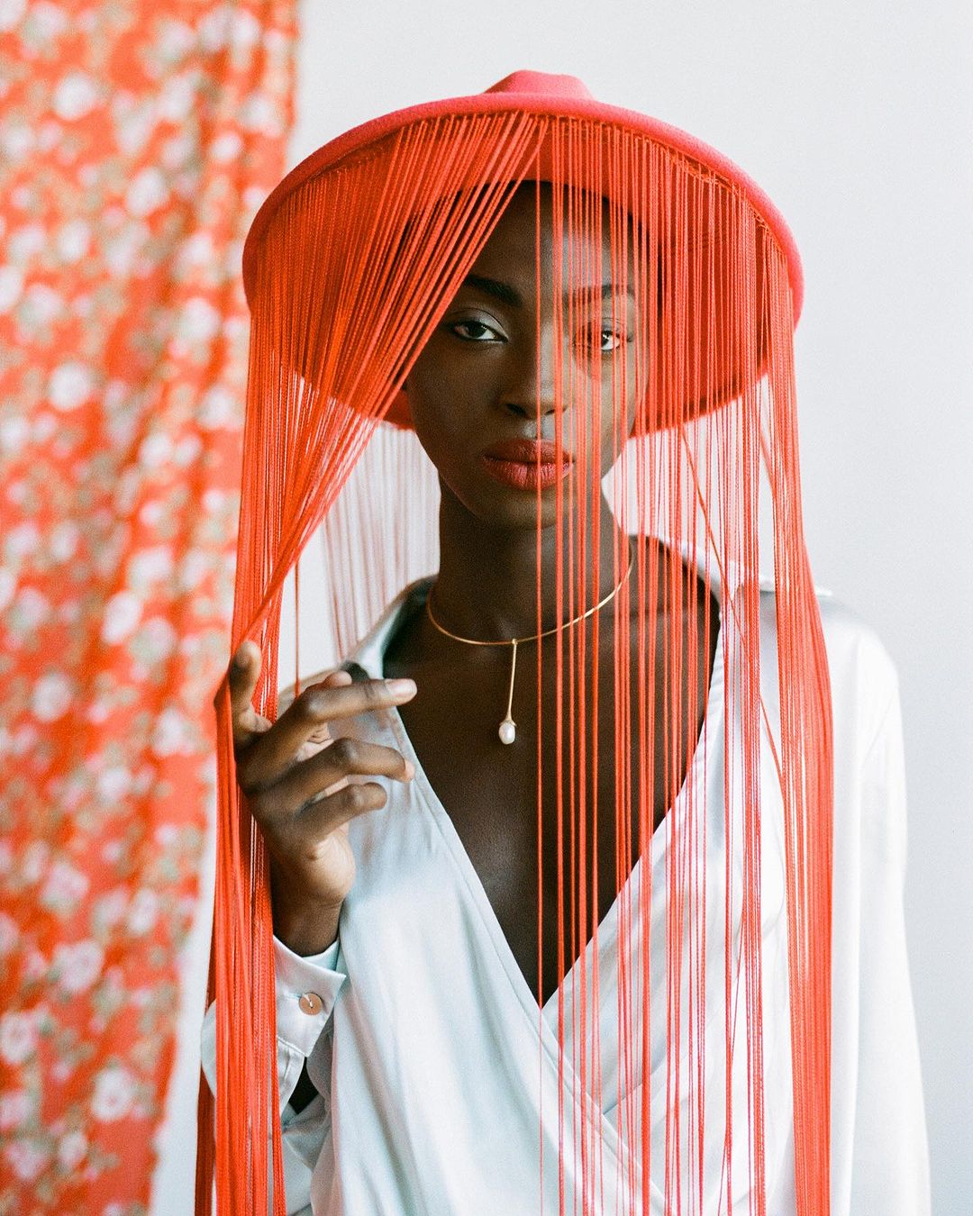 Portrait of an African American woman wearing a red hat with fringe, a white shirt, and a gold necklace against a floral backdrop.