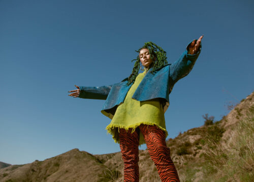 A young woman stands in an open field with arms outstretched, wearing a bright blue jacket, green top, and red tiger-striped pants against a blue sky.