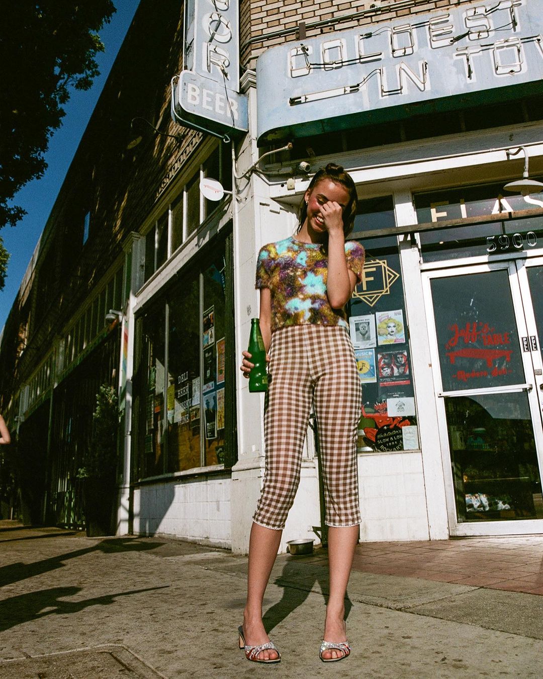 Young woman in patterned shirt and capri pants stands outside storefront holding a green bottle, casual and stylish in the city.