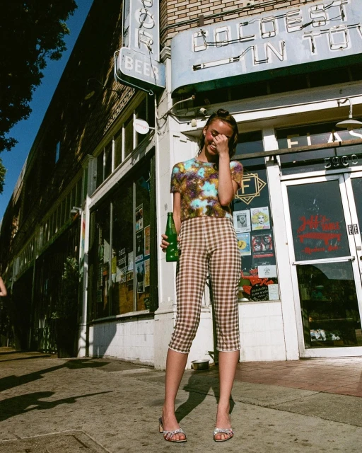 Young woman in patterned shirt and capri pants stands outside storefront holding a green bottle, casual and stylish in the city.