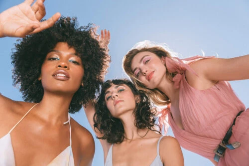 Three diverse women pose together in an eye-level shot against a bright blue sky, showcasing fashion and beauty styles, with hair and makeup.