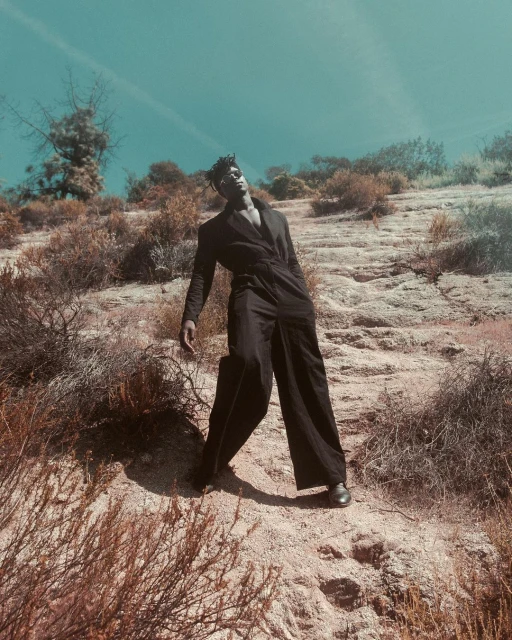 An African-American male model in a dark jumpsuit poses outdoors on a rocky hillside with scrub brush against a blue sky backdrop.