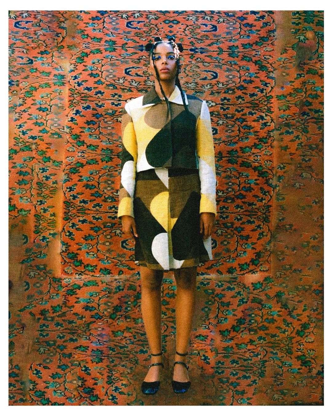 Full shot of woman in cropped jacket and mini skirt with an orange and green pattern background posing in front of the camera.