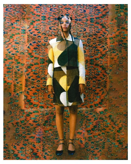 Full shot of woman in cropped jacket and mini skirt with an orange and green pattern background posing in front of the camera.