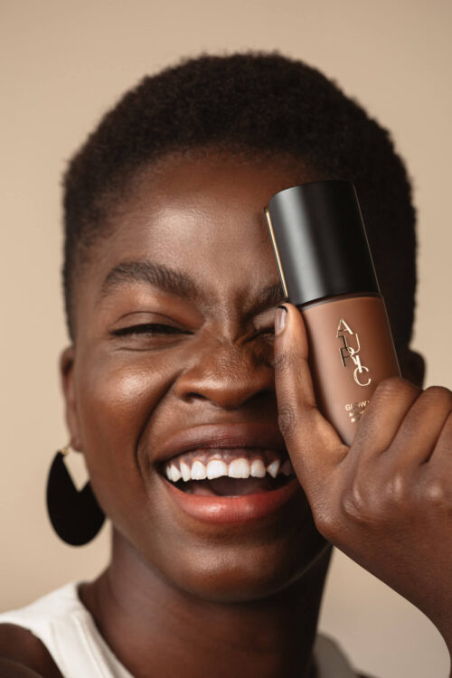 Smiling Black woman with dark hair holding up foundation bottle covering one eye in a beauty shot against a neutral background.