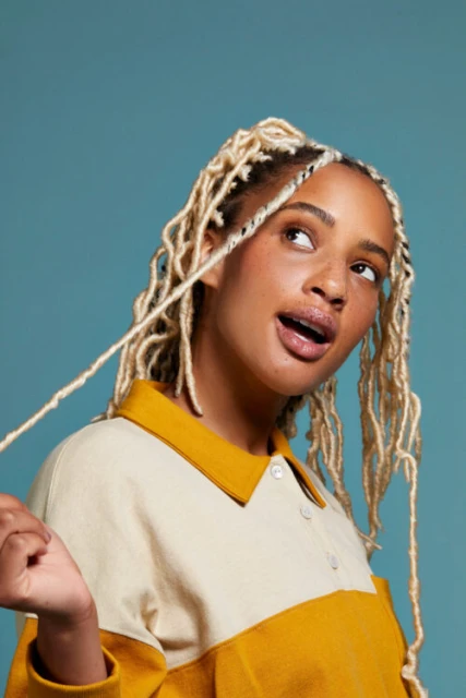 Close-up of an adult female with blonde braided hair and a mustard-and-cream collared shirt, looking upwards against a teal background.