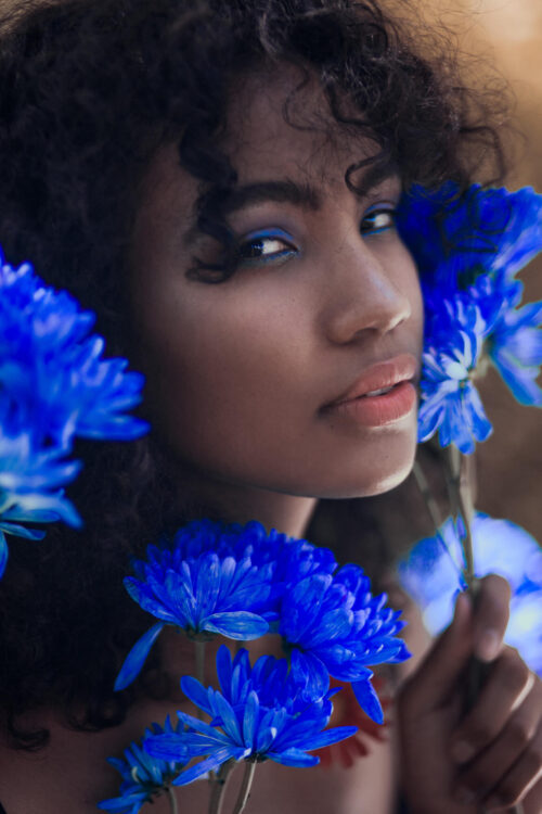Close-up portrait of a dark-skinned woman with curly hair, blue eye makeup, and surrounded by vibrant blue flowers against a soft background.