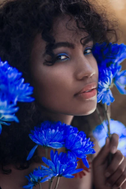 Close-up portrait of a dark-skinned woman with curly hair, blue eye makeup, and surrounded by vibrant blue flowers against a soft background.