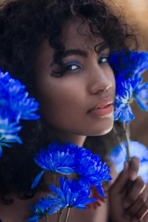 Close-up portrait of a dark-skinned woman with curly hair, blue eye makeup, and surrounded by vibrant blue flowers against a soft background.