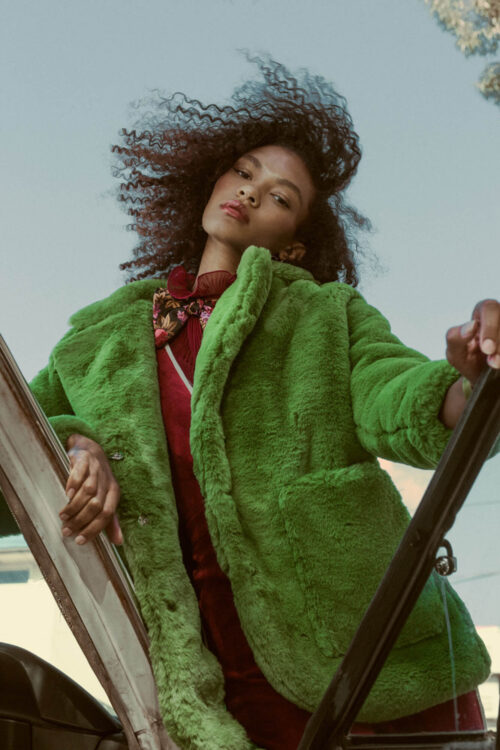 An adult woman with curly hair stands outside wearing a bright green faux fur coat, burgundy dress, and a floral print top. The sky is blue.
