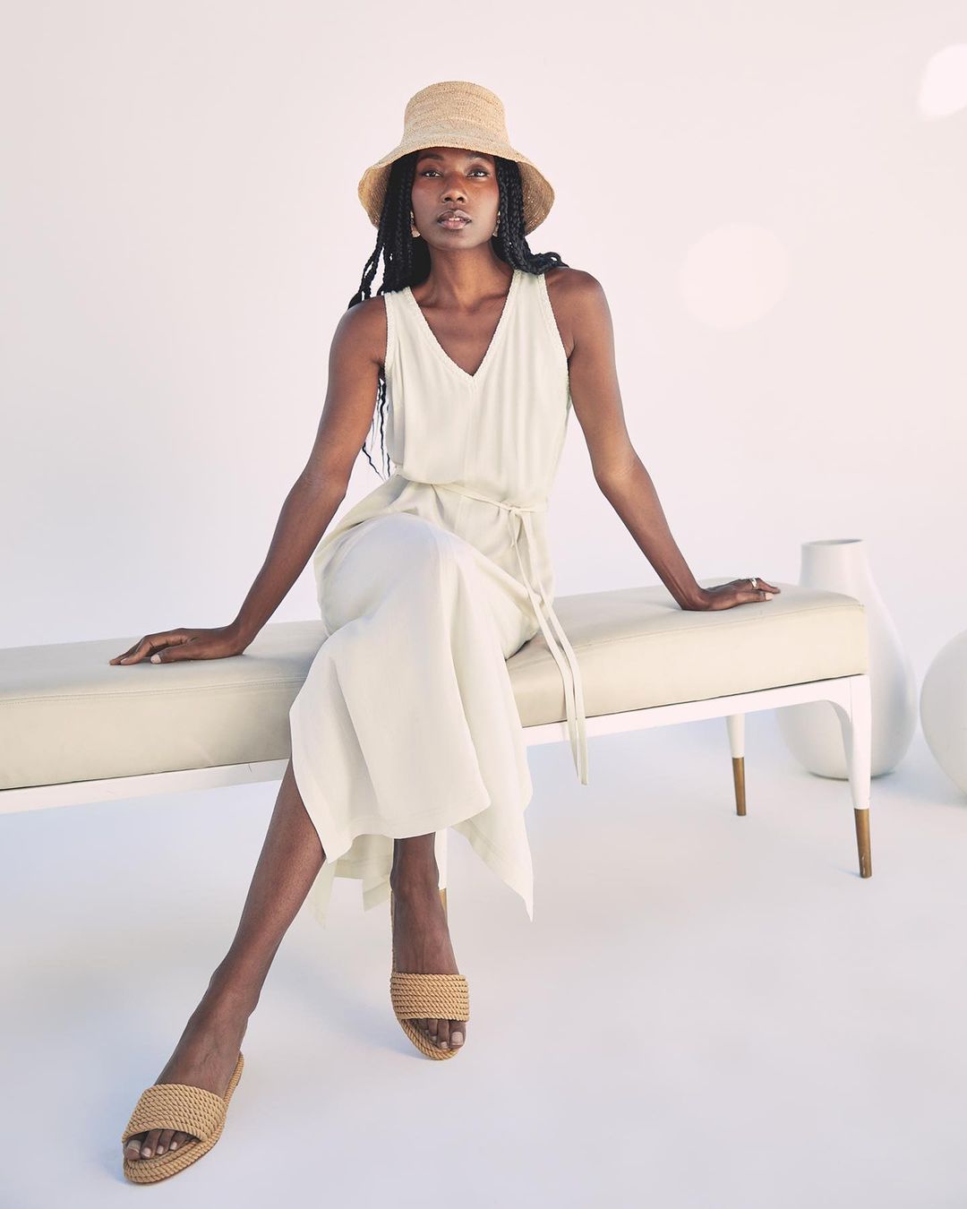 A stylish dark-skinned woman in a straw hat and cream dress sits on a bench in a bright studio with vases in the background.