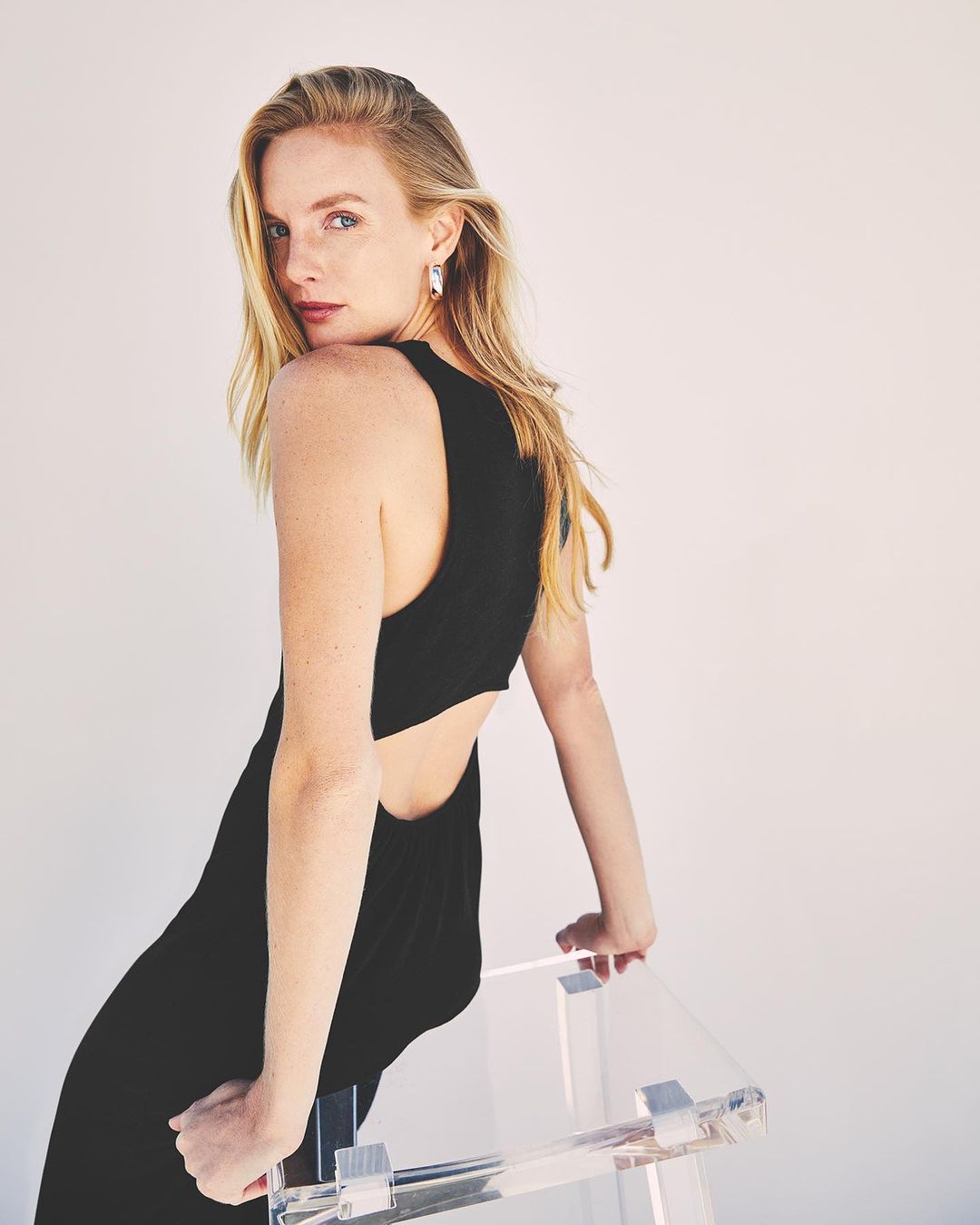 Blonde woman in black dress leans on acrylic chair in a bright, minimalist studio. Shoulder-length hair is styled, and she is looking back.