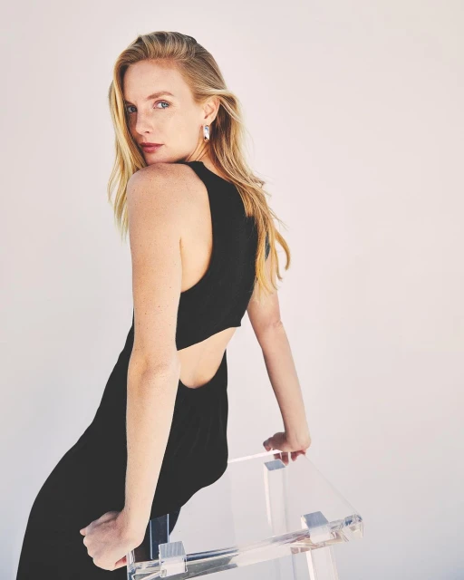 Blonde woman in black dress leans on acrylic chair in a bright, minimalist studio. Shoulder-length hair is styled, and she is looking back.