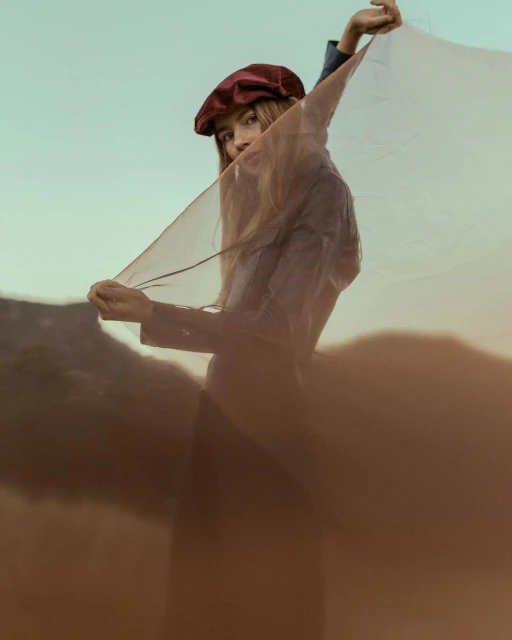 Portrait of a female model in a cap, holding a sheer fabric against a blurred mountain backdrop in a soft, dreamy outdoor setting.