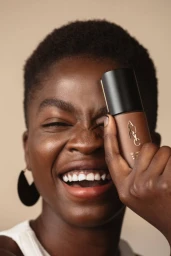 Close-up portrait of a Black woman smiling and holding a cosmetic bottle in front of her eye, photographed by Lauren Naylor.