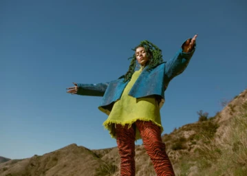 Low-angle outdoor portrait of a woman with green locs wearing a blue jacket, yellow dress, and red zebra-print trousers against a clear sky.