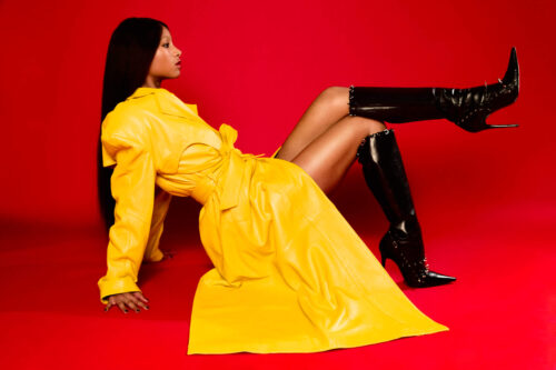 An adult female poses in a yellow trench coat and black, knee-high boots on a red backdrop in a fashion or beauty editorial photograph.