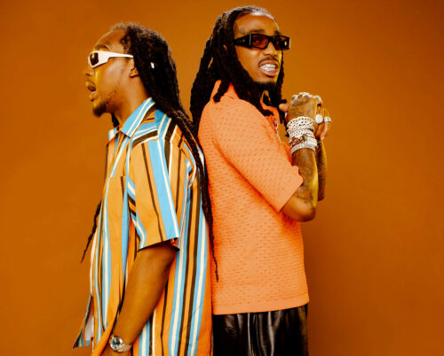 Two adult males with dreadlocks pose back-to-back in a studio setting, wearing stylish outfits and sunglasses against a brown backdrop.