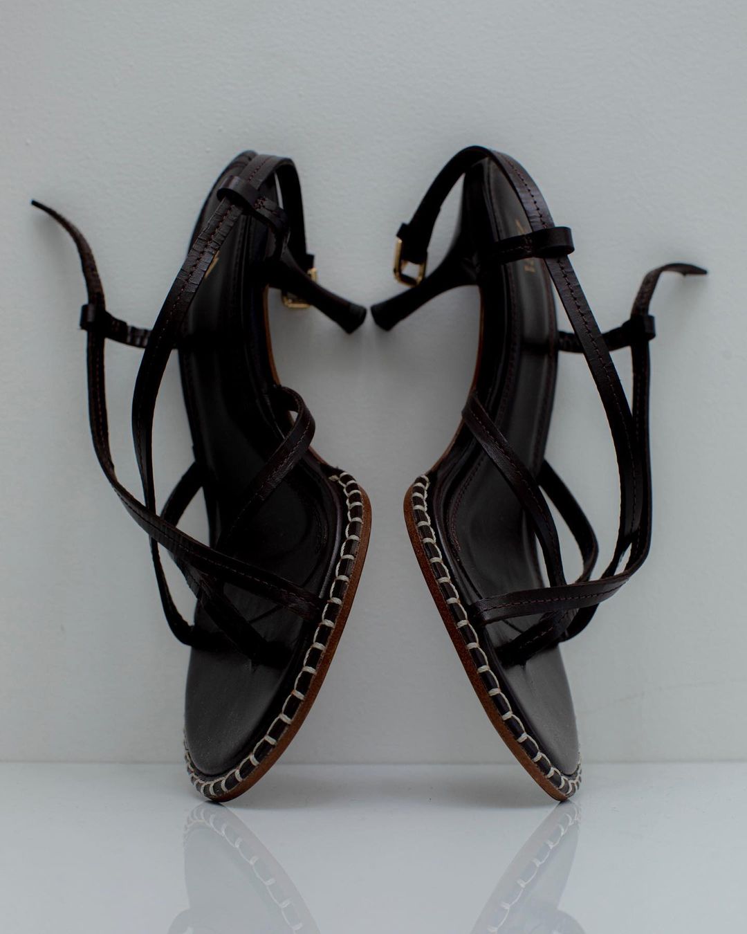 Elevated view of two dark brown strappy heeled sandals with ankle straps displayed on a white reflective surface.