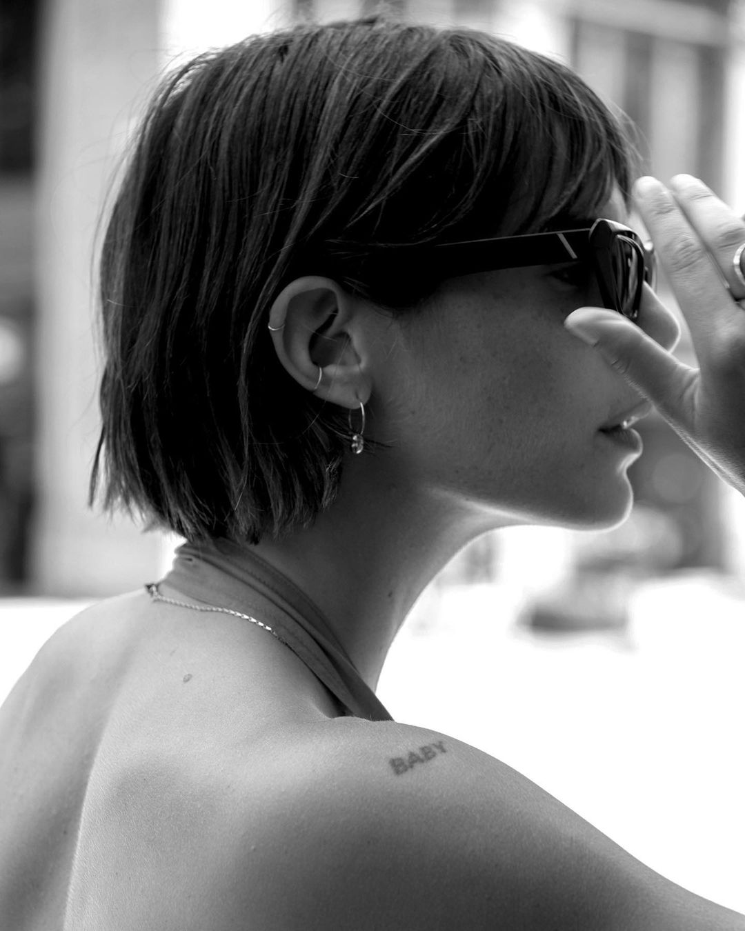Black-and-white portrait of an adult woman with short dark hair and sunglasses, captured in a side profile view with hand adjustment.