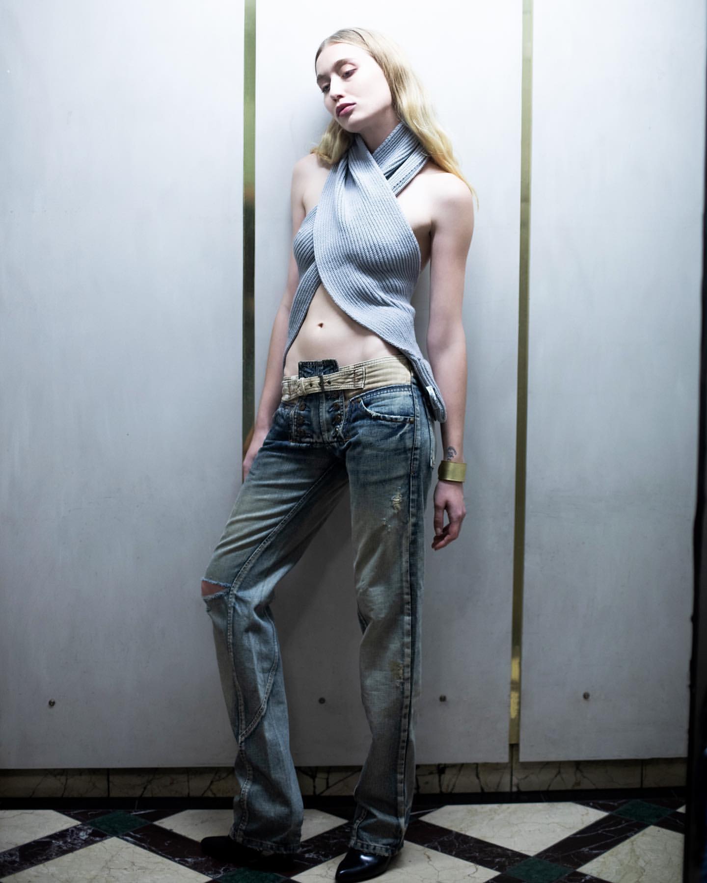 A blonde female fashion model poses in distressed jeans, a ribbed halter top, and black heels against a paneled wall with metallic trim.
