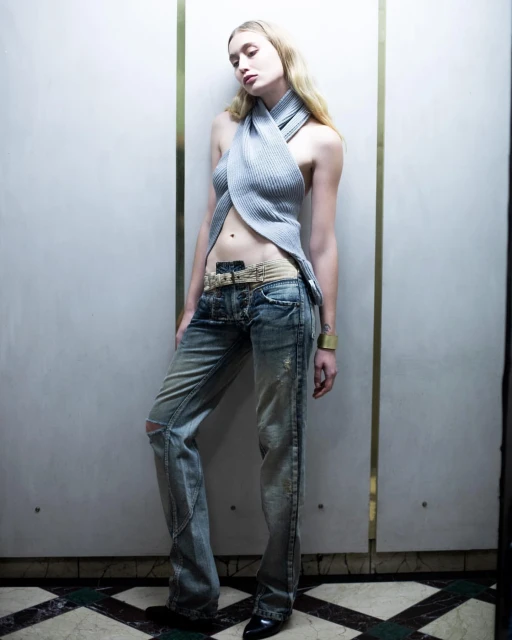 A blonde female fashion model poses in distressed jeans, a ribbed halter top, and black heels against a paneled wall with metallic trim.