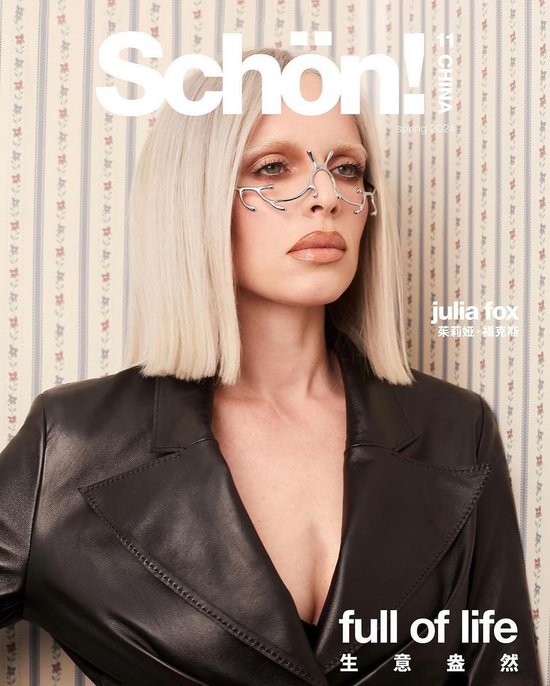 Julia Fox, a blonde woman with dramatic makeup and geometric glasses, poses in a black jacket in front of a floral wallpaper.