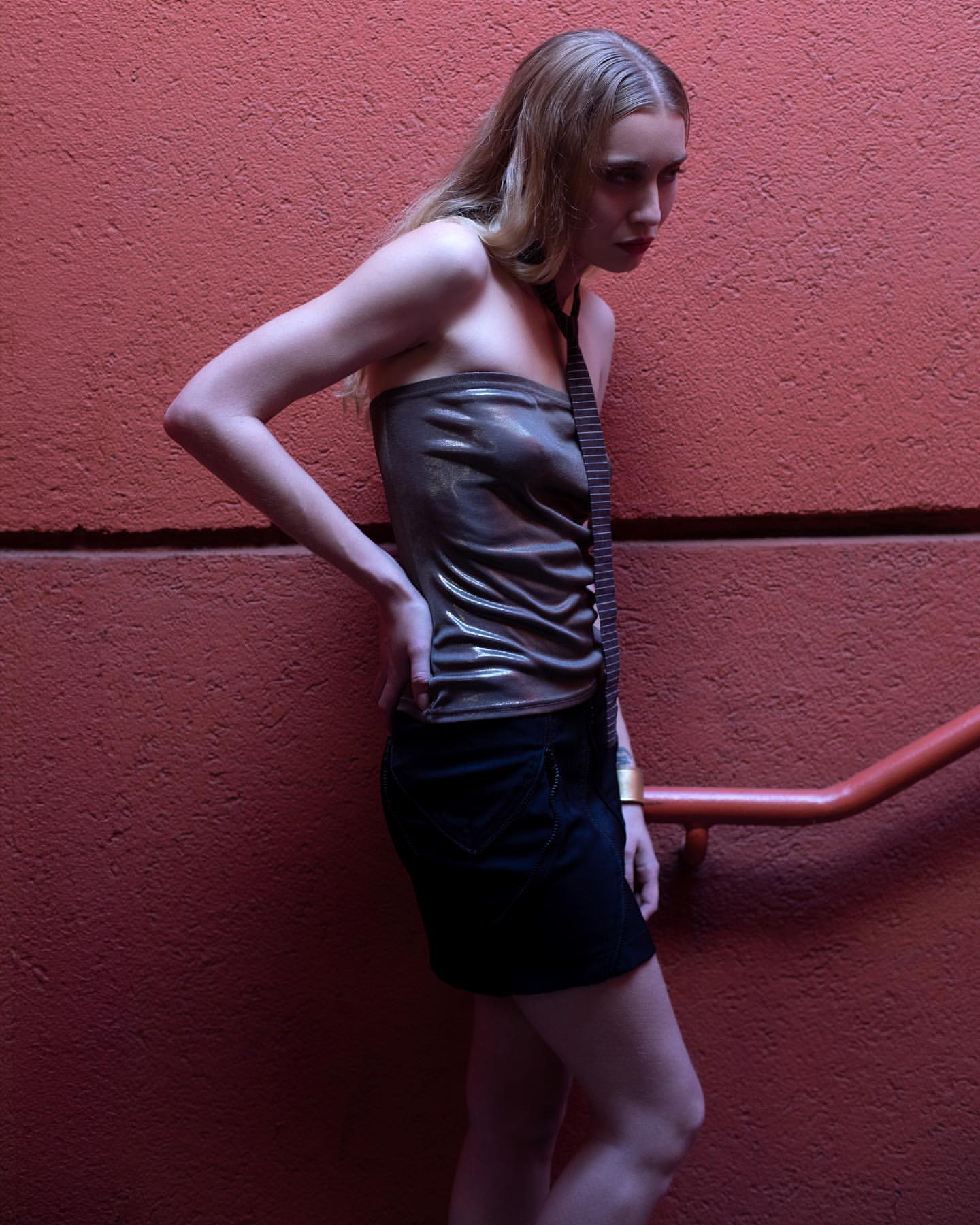 A slender woman in a metallic brown tube top and black miniskirt poses in front of a textured red wall, illuminated by soft lighting.