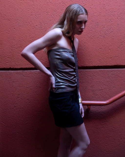 A slender woman in a metallic brown tube top and black miniskirt poses in front of a textured red wall, illuminated by soft lighting.