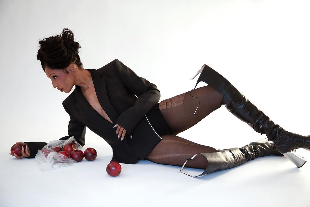 A woman in a black blazer and thigh-high boots poses with a bag of red apples in a bright, high-key studio environment. Fashion.