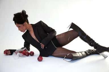 Model in black blazer and thigh-high boots reclining on a white background beside a plastic bag of red apples