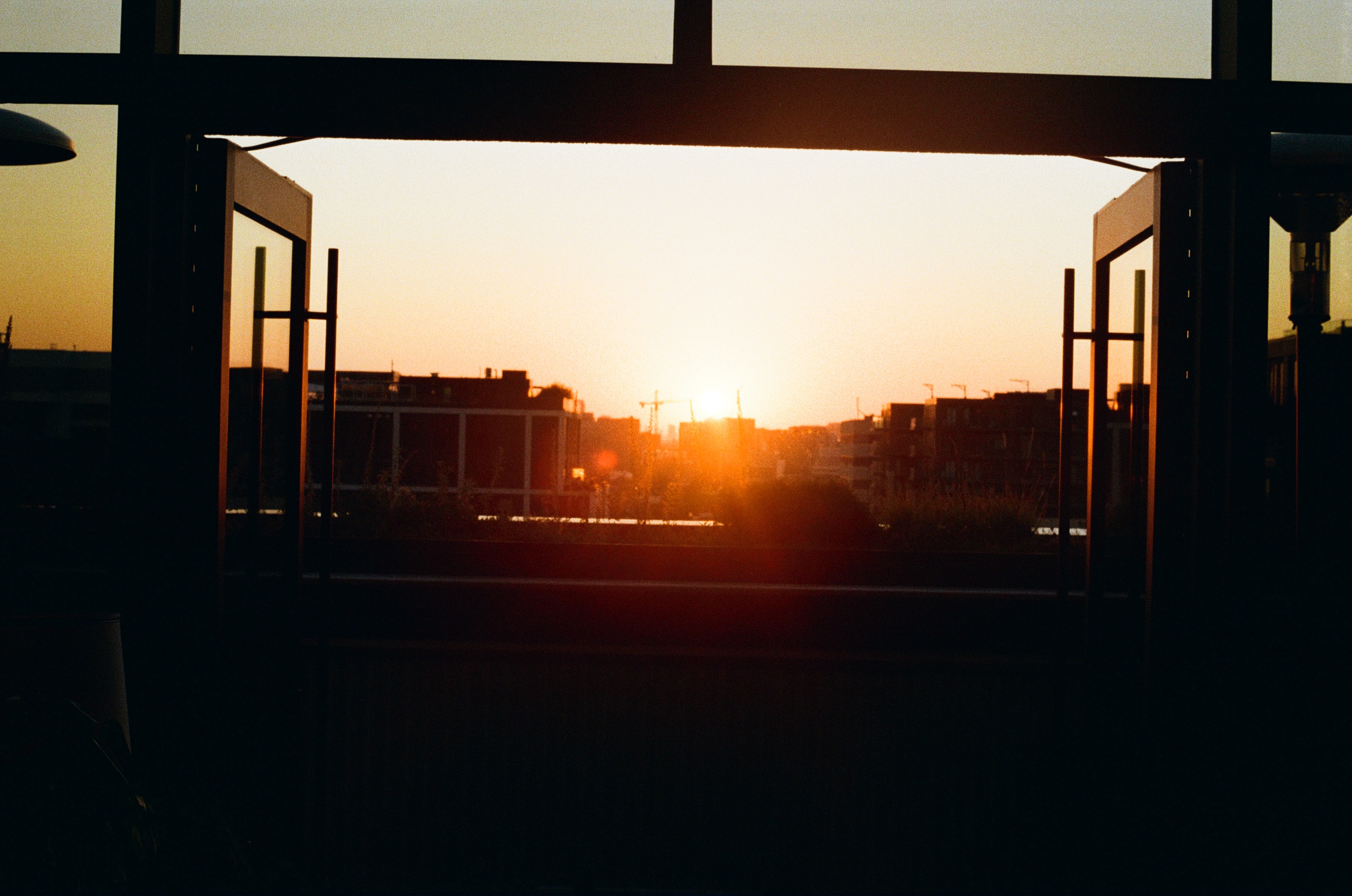 Scenic view of cityscape buildings at sunset, framed by architectural beams and windows, creating a dramatic and warm visual effect.