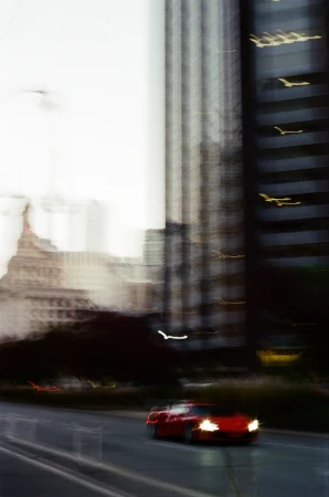 Red car races down a city street with tall buildings in the background, motion blur effect creates a sense of speed.