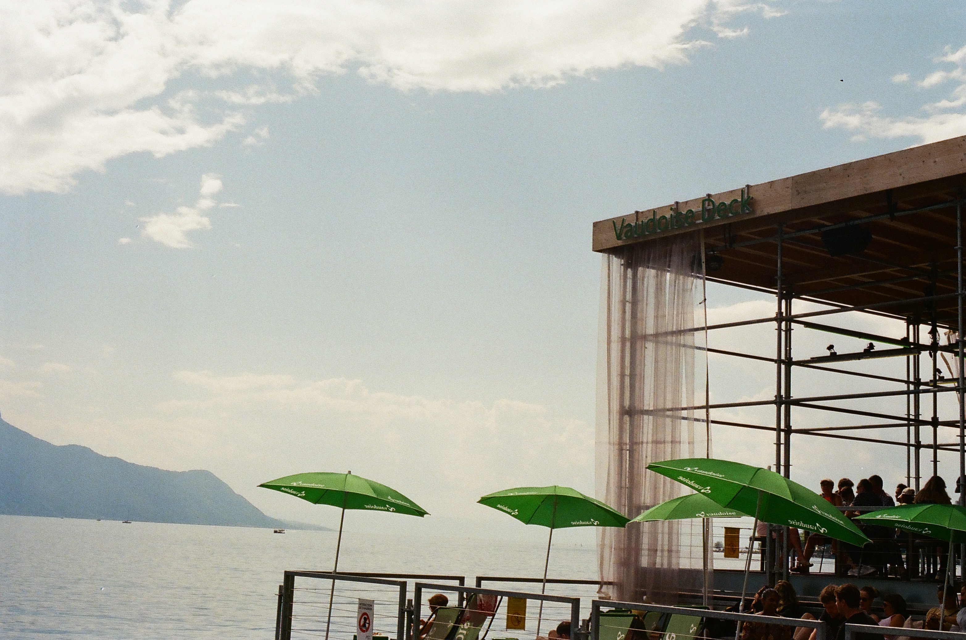 Green parasols and a platform structure on a sunny lakeside with mountains in the distance under a partly cloudy sky.
