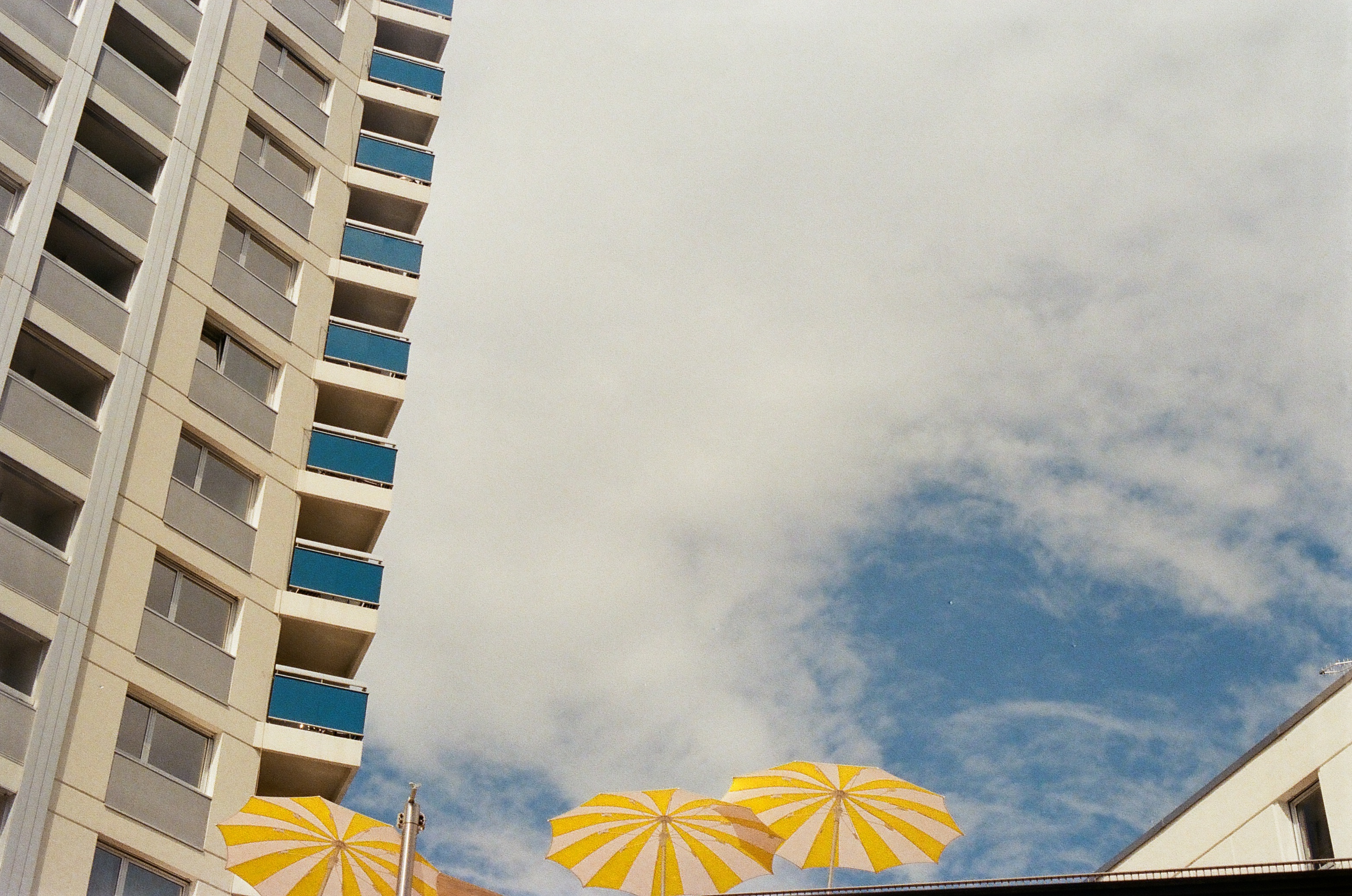 A low-angle shot showcases a modern apartment building beside yellow and white striped umbrellas against a partially cloudy sky.