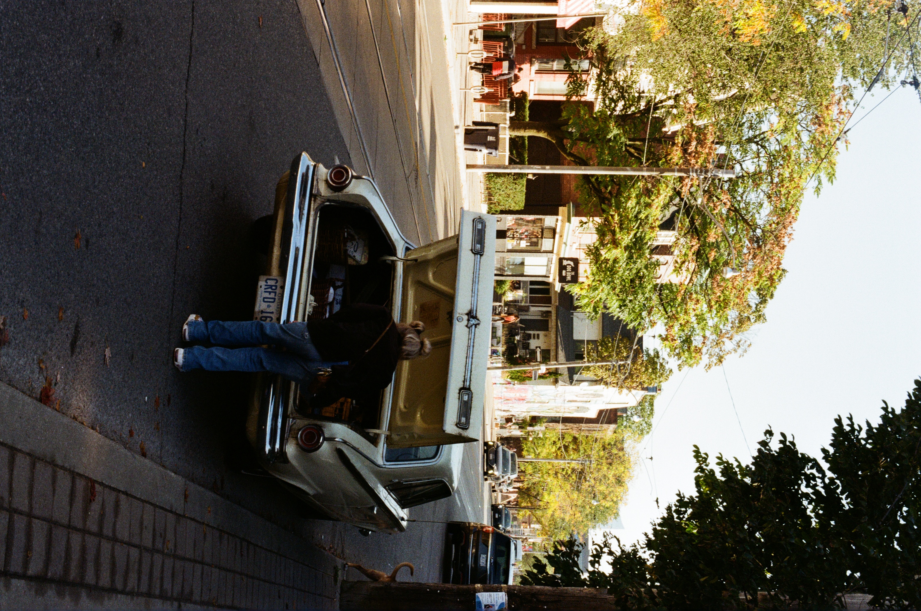A person stands at the trunk of a vintage car parked on a city street with buildings and trees in the background, captured in daylight.