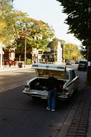 A person stands at the trunk of a vintage car parked on a city street with buildings and trees in the background, captured in daylight.