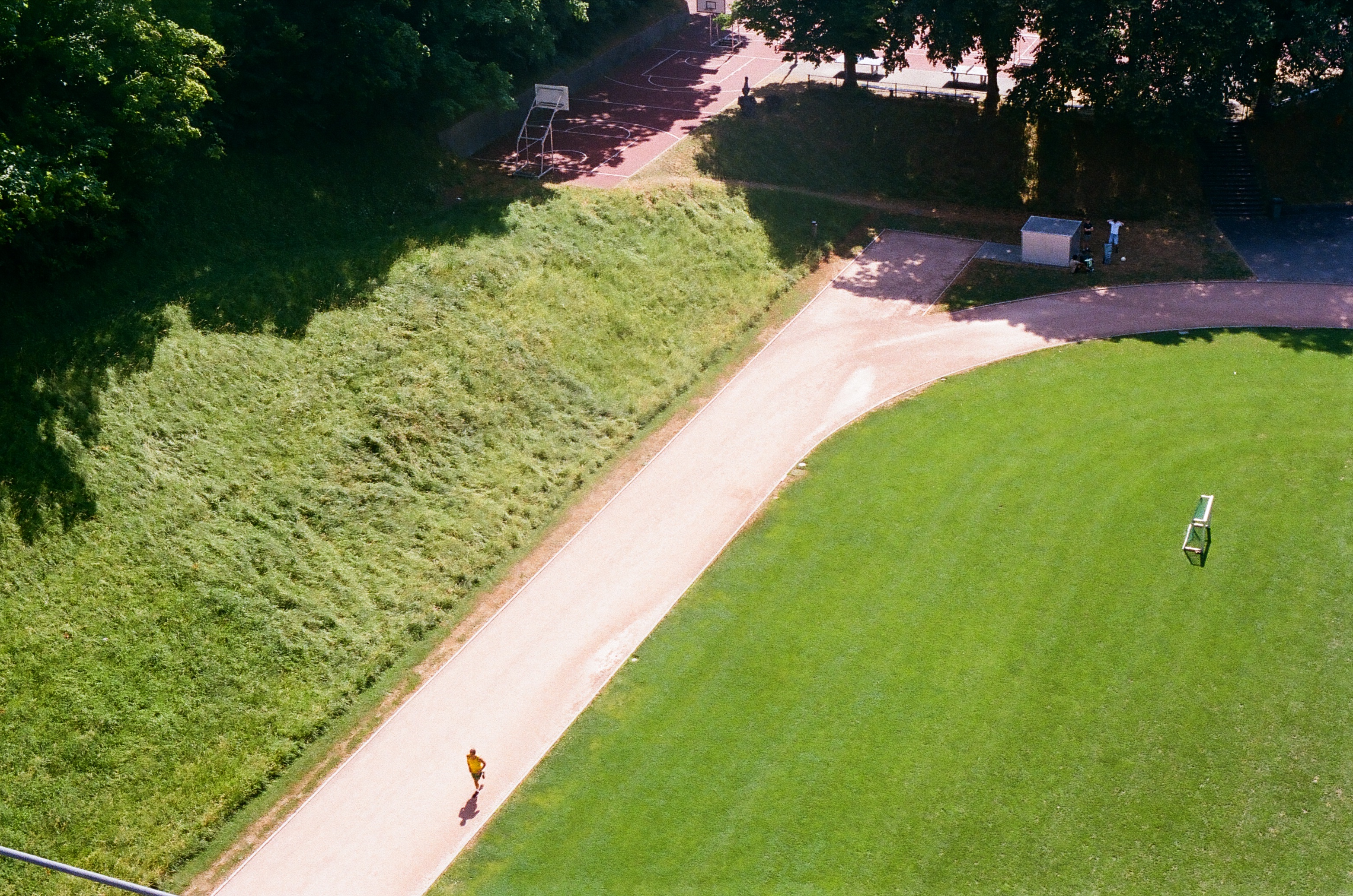 Aerial view of a runner on a tan path between a green lawn and grassy hill with a basketball court in the background on a sunny day.