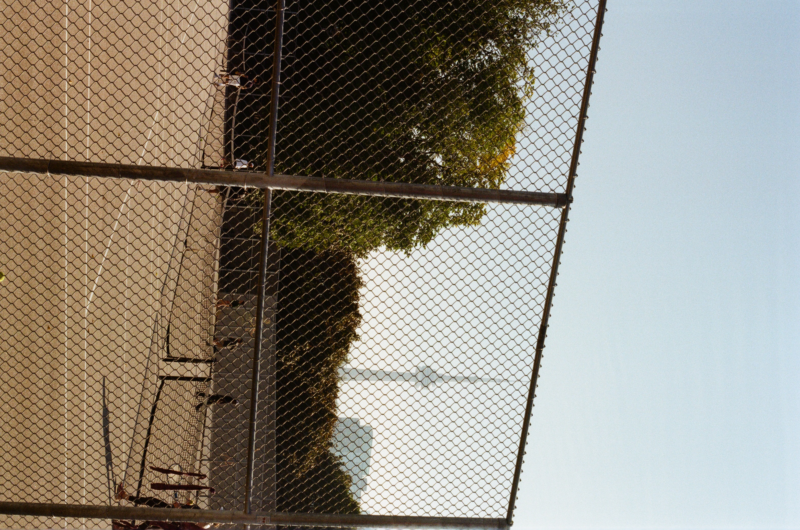 Tennis game in progress on outdoor court viewed through chain-link fence; distant skyline includes a tall communications tower.
