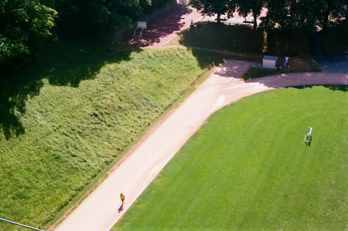 High-angle view of a solitary runner in a yellow shirt exercising along a curved outdoor track next to a green lawn.
