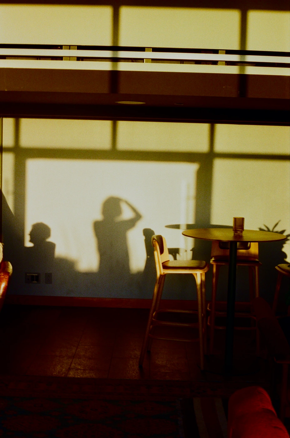 Dramatic shadows of people projected onto a sunlit wall beside a wooden high-top table and barstools in a warm interior.