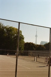 A wire chain-link fence in the foreground looking onto an outdoor tennis court with distant trees and a tower on the horizon.