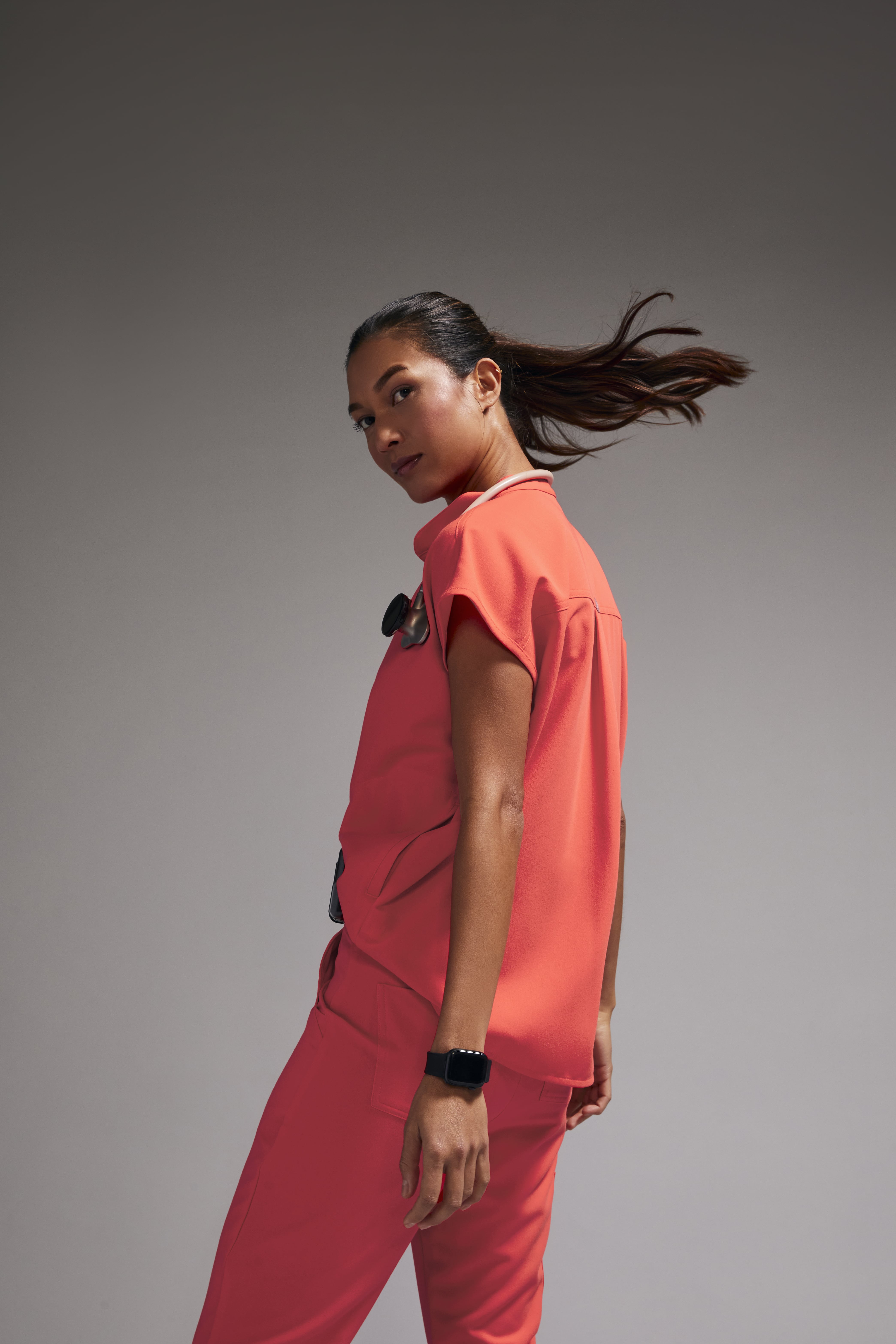 A young woman in coral scrubs and a stethoscope looks over her shoulder in a studio portrait against a neutral gray background.
