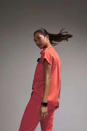 A young woman in coral scrubs and a stethoscope looks over her shoulder in a studio portrait against a neutral gray background.